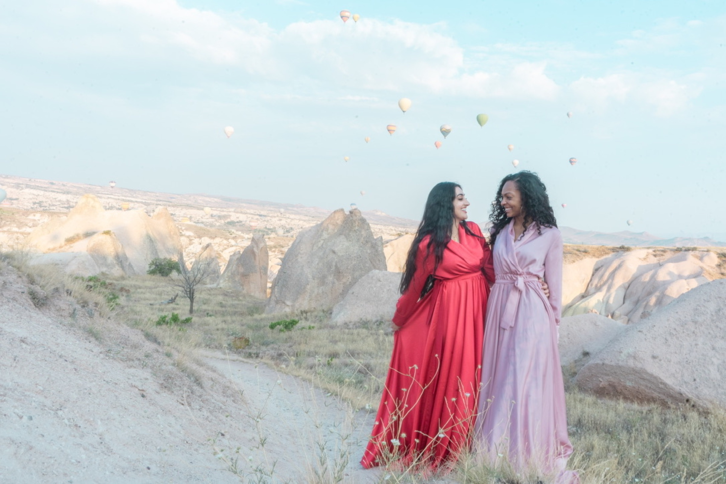 Two women smiling together in a valley with hot air balloons behind them