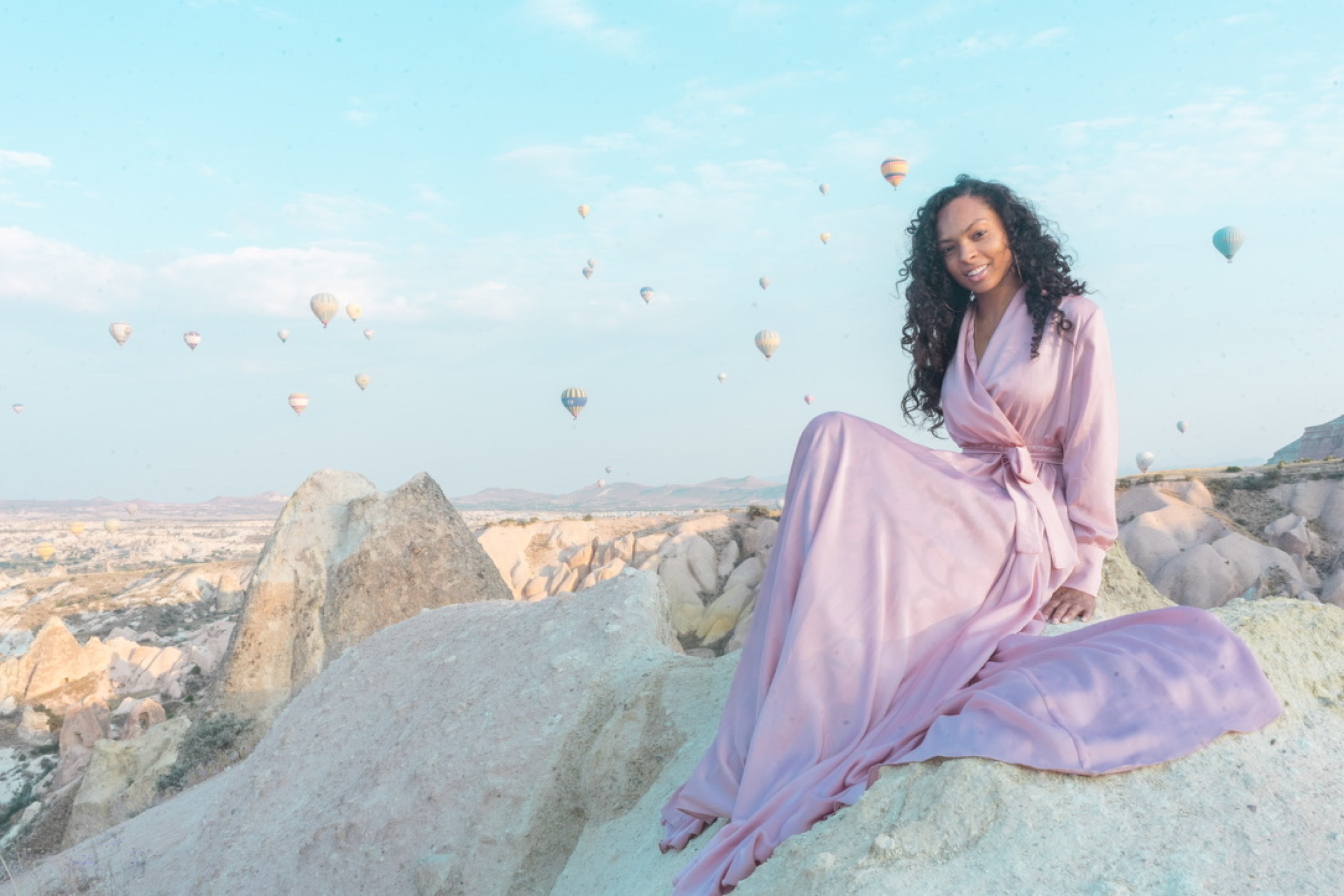 A woman in a rose-colored gown seated on rock formations with balloons in the sky
